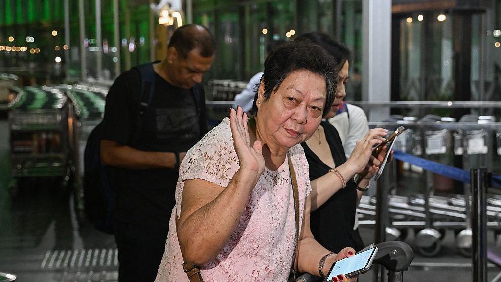 A middle-aged woman with short black hair in a pink shirt waves as she looks to the side, at the entrance of an airport