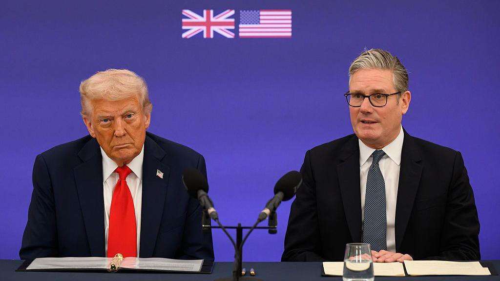 Trump dhe Starmer janë ulur në një tavolinë të zezë me mikrofonë të zinj. Një ekran me ngjyrë vjollcë pas tyre ka flamuj të vegjël të Mbretërisë së Bashkuar dhe SHBA-së të shtypur në të. Starmer ka veshur një kostum të zi dhe kravatë me motive me një këmishë të bardhë. Starmer shikon përpara me duart e kryqëzuara mbi njëra-tjetrën. Trump shikon drejt përpara dhe duket i vrenjtur. Ai ka veshur një kostum blu të errët me kravatë të kuqe dhe një këmishë të bardhë.