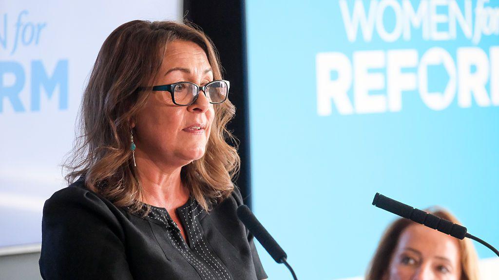 Linden Kemkaran wearing glasses and talking into two podium microphones. Light blue and white backgrounds can be seen behind her.