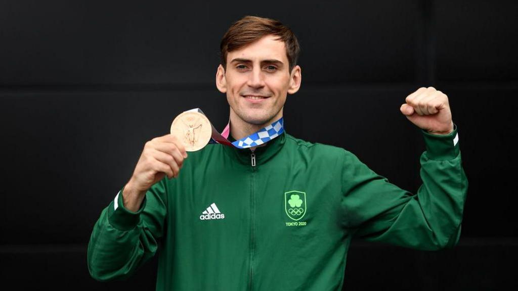 A man with short brown hair is smiling at the camera. He is holding one arm up and making a fist, in the other he is holding an Olympic medal. He is dressed in a green zip jacked with 'Adidas' and 'Tokyo 2020' branding. 