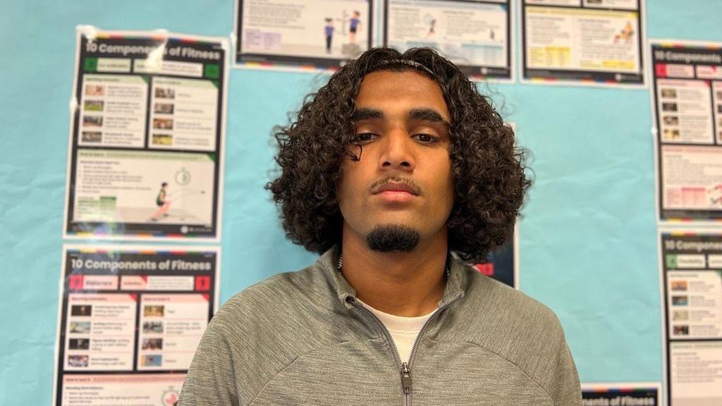 Mohammed is behind a classroom that has lots of different leaflets he is wearing a grey zipped up jacket.