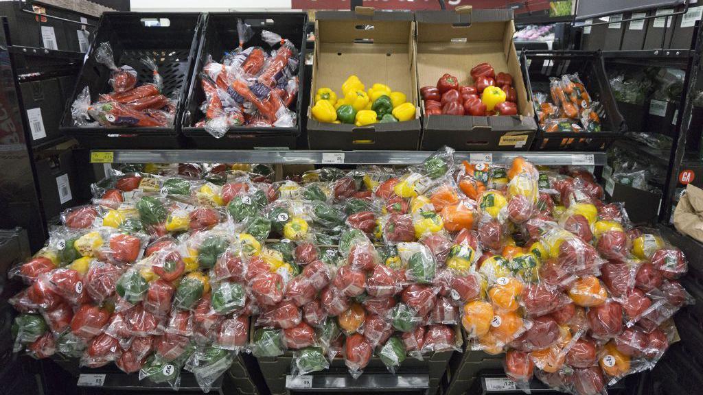 Packets of green, yellow and red peppers are laid out in a super market, with loose one above them in a cardboard box.