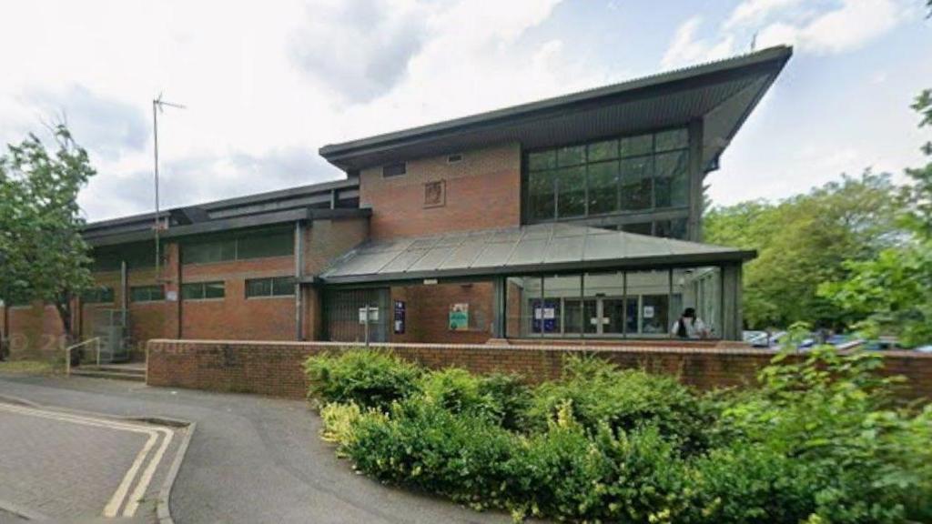 A building in the background, with a road in the foreground. There are bushes by a wall by the pavement.