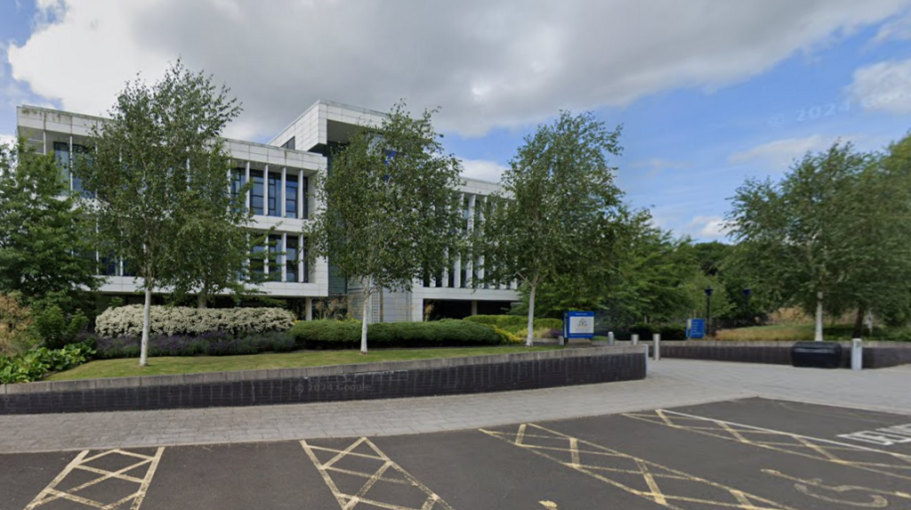 The Durham Constabulary headquarters building sits behind large trees. It is a white clad, long, flat-roofed, three-storey modern effort with large windows set behind thin pillars. A car park is in front of the building.