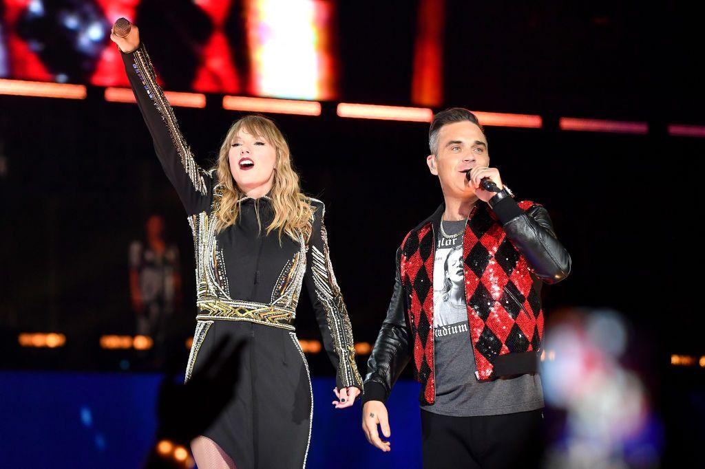 Taylor Swift and Robbie Williams on stage together in London in 2018. She wears a black dress with metallic details and he wears a Taylor Swift Reputation top with a red and black sparkly jacket on top. 