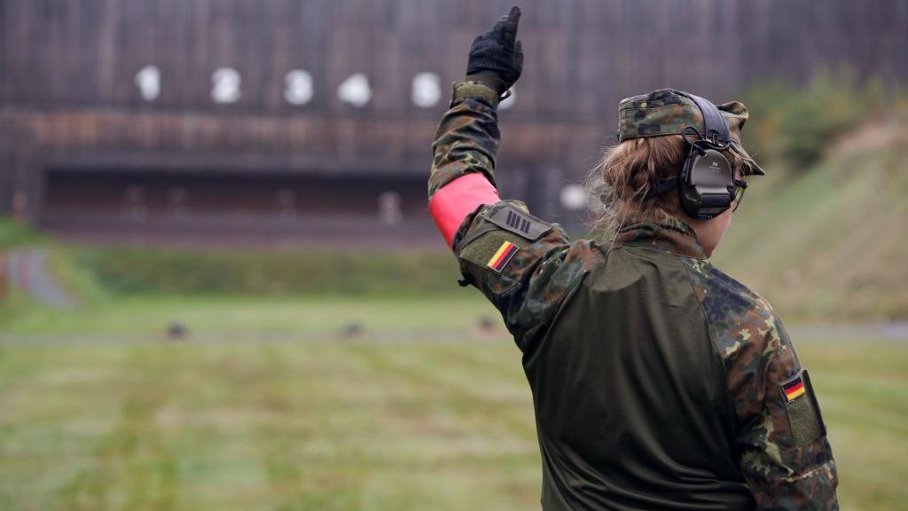 A German drill instructor at a firing range holds up her left arm