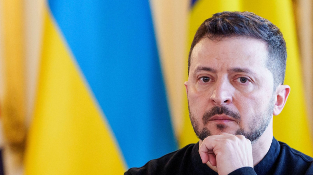 Close up of Volodymyr Zelensky sitting down in black shirt, a wall of Ukrainian blue and yellow flags behind him