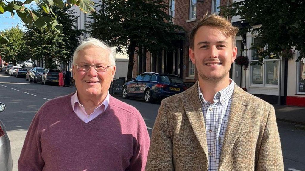 Gareth Morgan (left) stands next to Glyn Preston (right) in a street. They both smile at the camera in the sunshine. Mr Morgan wears a purple v-neck jumper and pink shirt, he has silver short hair and wears thin rimmed sunglasses. Mr Preston wears a tweed suit jacket and a white checked shirt and has light brown hair.