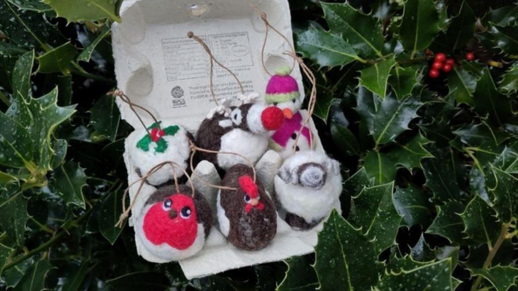 Six felt Christmas decorations. They include snowmen, robins and a Christmas pudding. They are displayed in an egg box which has been positioned among holly leaves.