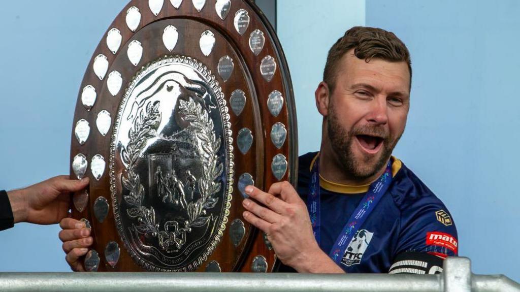 Scott Arnold holding a shield trophy and celebrating wearing his football kit and the captain's armband