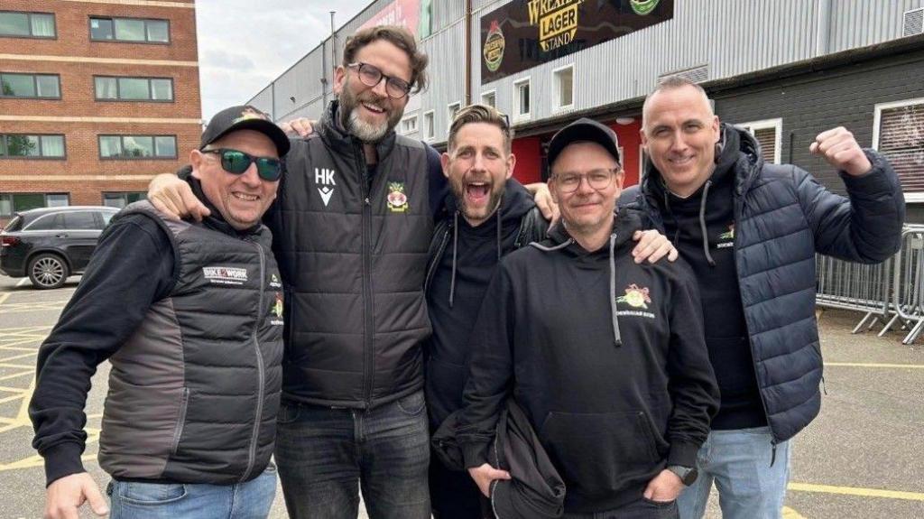 Norwegian fans smile and celebrate outside the Racecourse football ground with Wrexham AFC director Humphrey Ker