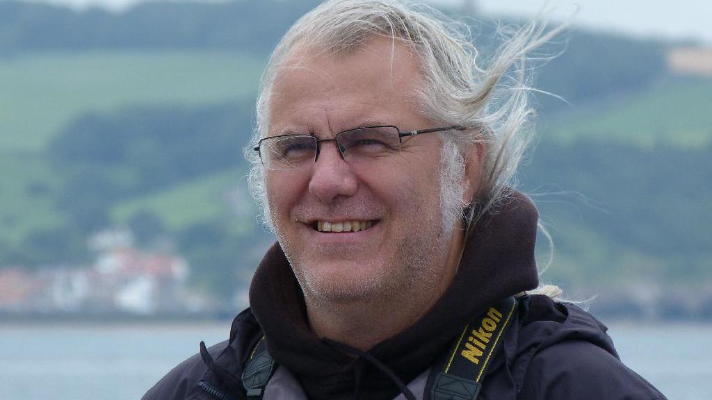 A smiling Robin Petch has long grey hair and wears a black jacket and  dark metal-framed spectacles. He looks windswept in front of a backdrop of the sea and green hills.