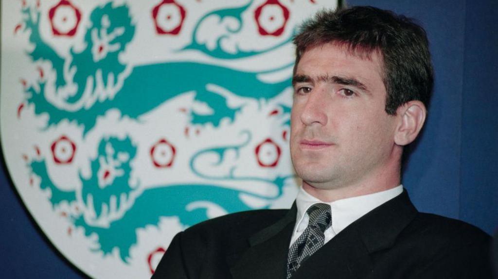 Dark haired man in dark suit sits back on a chair with an England football crest in the background