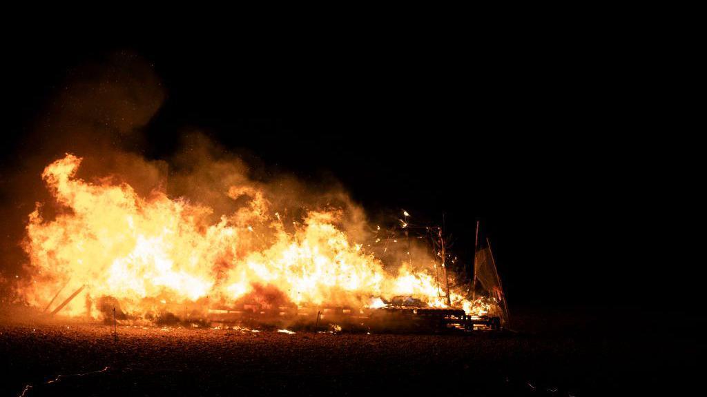 A large fire burns in the distance with the remnants of sticks and lanterns sticking up out of it. It is otherwise dark around the fire, which is just lighting up the stones of the beach.