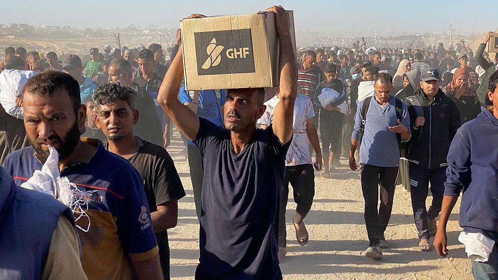 A photograph of a Palestinian man in Gaza looking towards the camera while holding a cardboard box with GHF branding above his head