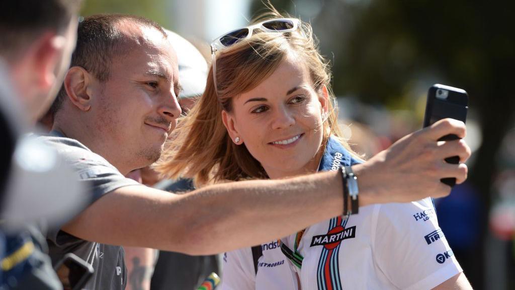 Susie Wolff in white team shirt with blue collar smiling as a fan in a grey T-shirt takes a selfie of them together. They are surrounded by other racing fans.