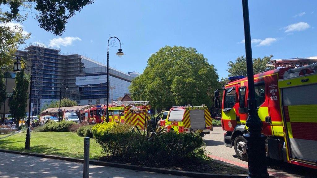 A number of fire engines and emergency vehicles parked in the road outside a tall block of flats. Other buildings and lamposts can be seen in the background.
