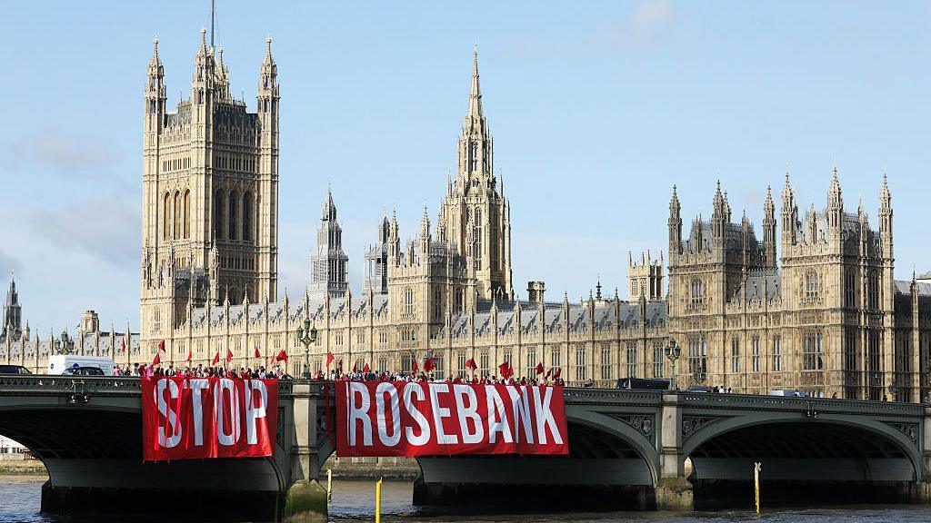A large red sign with white letters spelling out STOP ROSEBANK being hung of the side of a bridge in front of Westminster