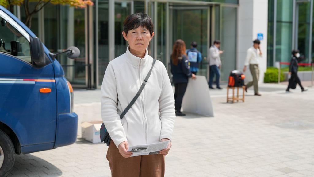 A middle-aged woman with short, dark hair wearing a white fleece stands outside a building