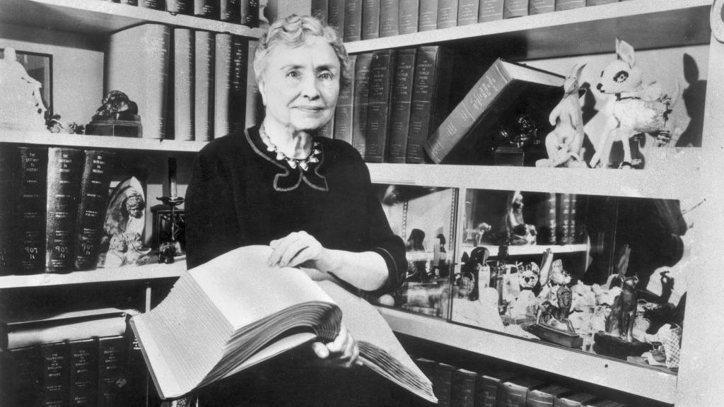 Helen Keller holding a Braille volume and surrounded by shelves containing books and decorative figurines. 