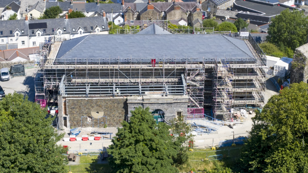 The gaol building undergoing construction across three floors is surrounded by green trees and houses.