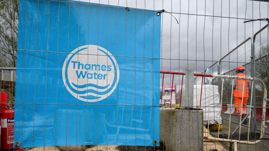 A fence with workers in the background with a blue Thames Water sign attached to it. In the background is a person in fluorescent orange work clothes.