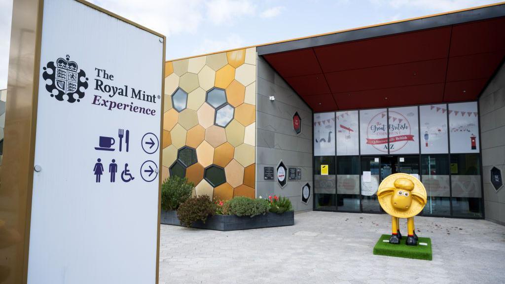 A general view of the Royal Mint in Llantrisant. There is a gold pattern on the wall of the building. The rest of the building is grey with glass windows and doors that lead to the entrance. A gold Shaun the sheep sits outside the building. A sign to the left of the picture says 'The Royal Mint Experience' with symbols of a cup and knife and fork to signal a cafe. It also has symbols of the toilets below. 