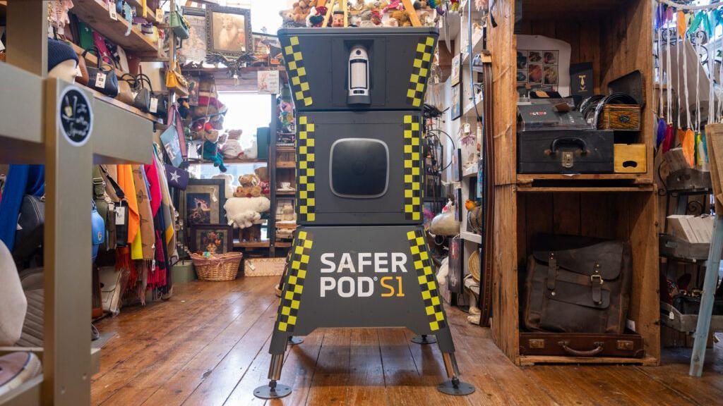 A black tall object in a shop surrounded by clothes and things. The object is called a Pod and has police white and black tape down the side of it. It is a three layer pod. The bottom layer as white writing saying safer pod. The second layer has a black square in the middle and the top layer has a white long light.