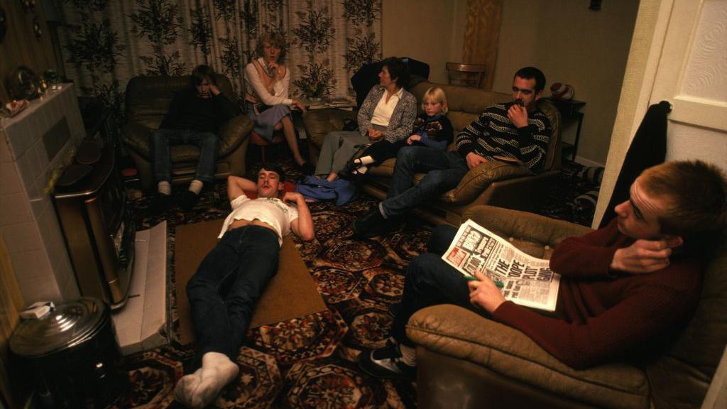 A photo of people sat in a living room in the 80s. There is a man laying on the floor with white socks, blue jeans and a white t-shirt. A man is on an brown armchair in the right of the photo, his right hand touches his left ear and he is holding a newspaper. He has a red jumper on and blue jeans. To the left of him are three people sat on a sofa. The one on the far left has a blue and grey striped jumper and jeans and has his hand on his mouth. In the middle is a child with blonde hair and next to them is a woman looking to her left with a blue cardigan, white shirt and blue trousers. To the right of her is a woman on a chair with a white top, peach apron and blue skirt, sitting cross-legged and to the right of her is a man on another brown armchair. He is blue jeans and a maroon jumper on and his hand on his chin. There is a fire place to the right of him. The carpet is patterned. 