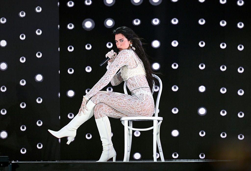 Dua Lipa dressed in a white lace bodysuit performs a song while sitting on a chair