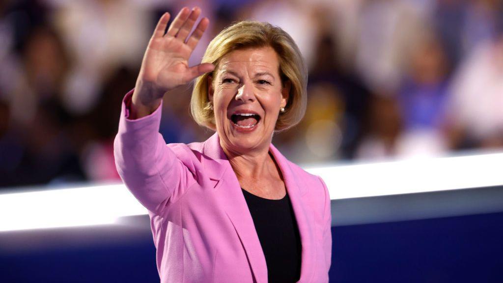 Tammy Baldwin, wearing a pink blazer over a black dress, gestures with her face and waves to the audience at the 2024 Democratic National Convention