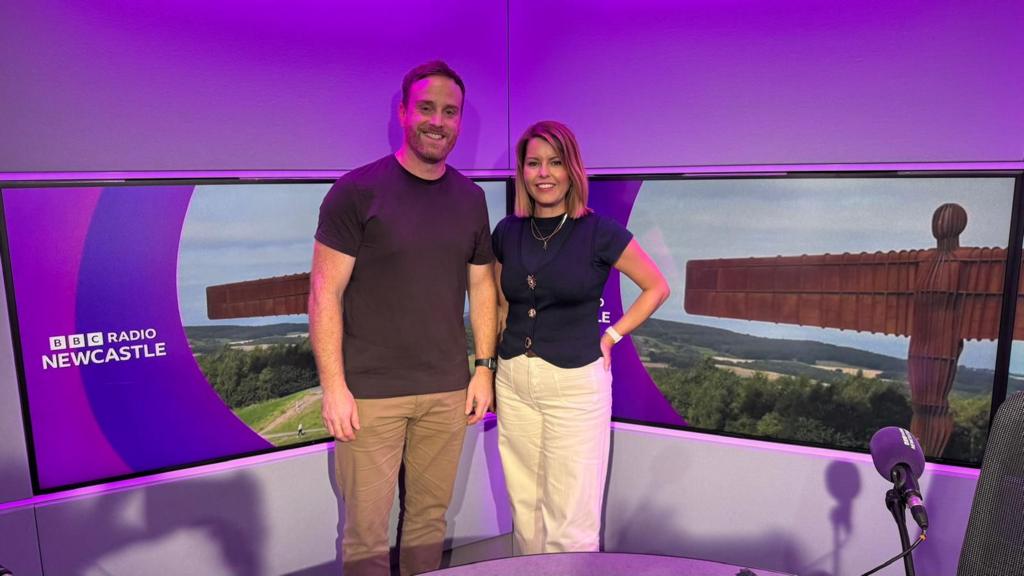 BBC Newcastle Breakfast presenter Matt Bailey stands next to North East Mayor Kim McGuinness. He is wearing a black t-shirt and light brown trousers and has short dark hair and stubble and she is wearing a blue short-sleeved top with three large brown buttons and has shoulder length light brown hair. They are standing in the BBC Newcastle studio which has purple on the walls and a large horizontal screen with the BBC Radio Newcastle logo in white lettering against a purple background and a photo of the Angel of the North