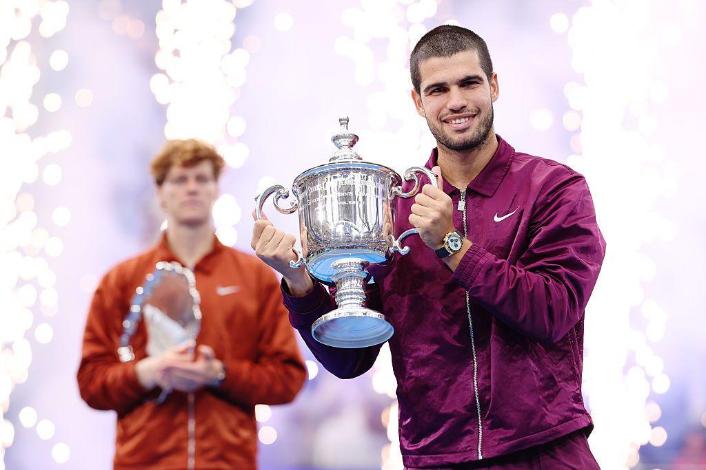 Carlos Alcaraz smiles while holding the US open trophy while Jannik Sinner stands behind him as fireworks erupt