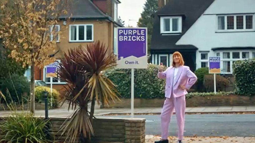 Still taken from an advert, showing a woman standing on a suburban pavement in front of 1930s homes. There are Purplebricks for sale signs in front of three houses. She is wearing a pink suit and has her thumb up.