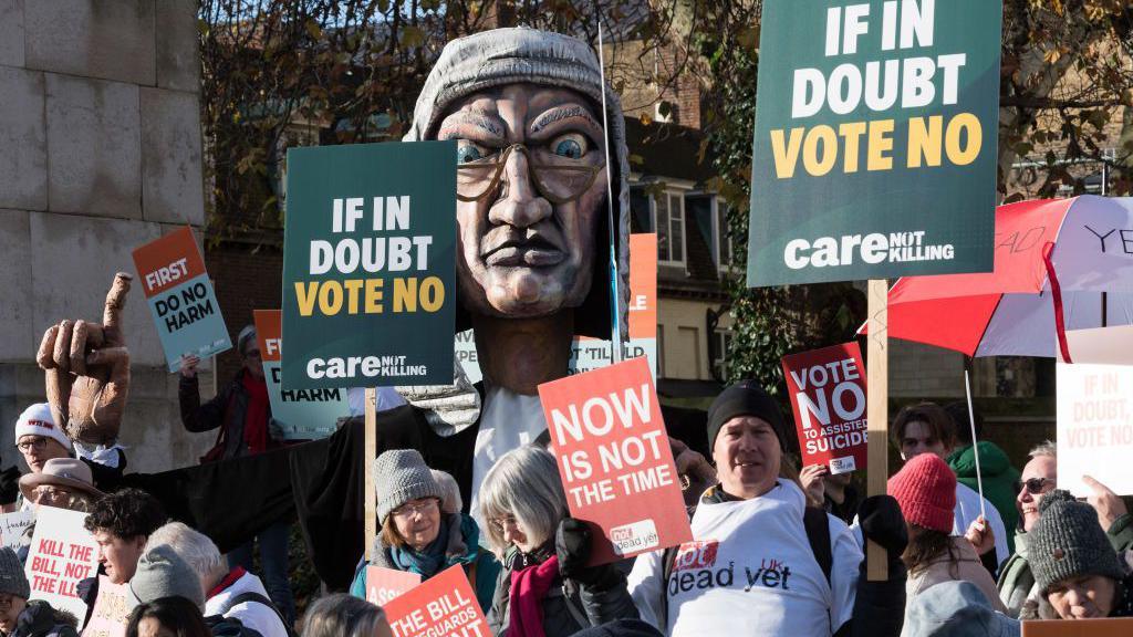 Campaigners opposing assisted dying protest outside Parliament, in front of a big puppet of a judge. They hold signs holding slogans including "if in doubt vote no", "now is not the time" and "first do no harm".