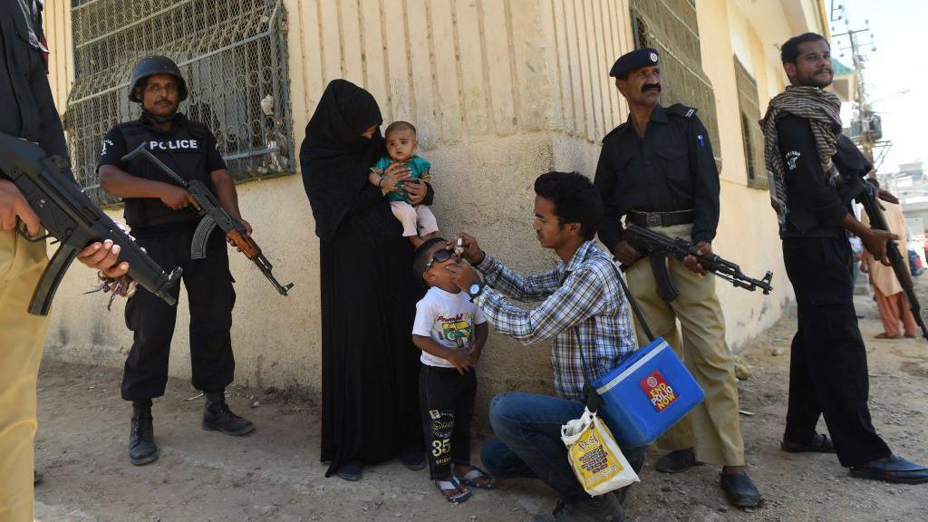 Pakistani policemen with guns stand guard, as a health worker administers polio drops to a small boy during a polio vaccination campaign in Karachi. 