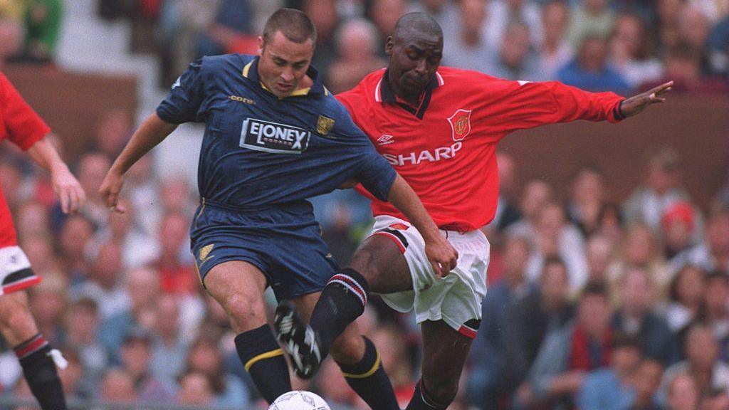 Gary Elkins is pictured playing for Wimbledon against Manchester United in the mid-1990s. He wears a blue football kit and is battling for the ball with Andy Cole who wears a red Manchester United shirt and white shorts.