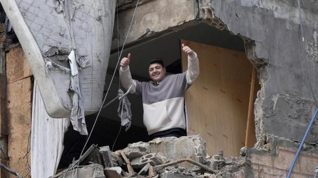 A man gives double thumbs up to the camera, smiling, framed by an enormous hole in a building