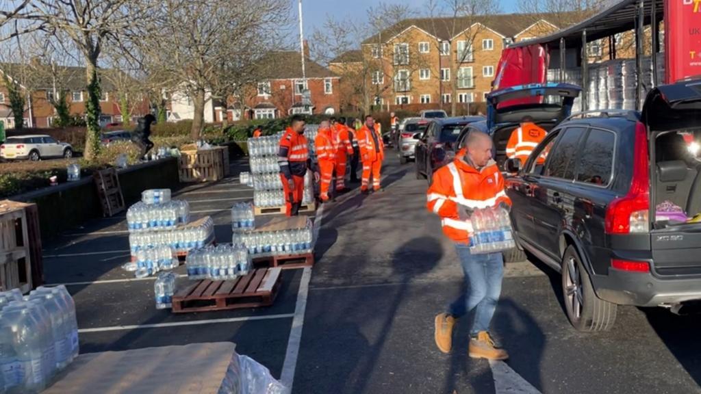 Water from a lorry is loaded into cars by men wearing orange jackets