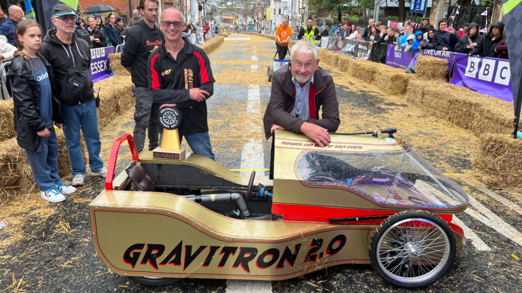 A gold kart is sideways on a road with two people standing and leaning on it. It is sat on the top of a road which has hay bales lined up either side and a silver fence. The kart says GRAVITRON 2.0 in black on the side. 