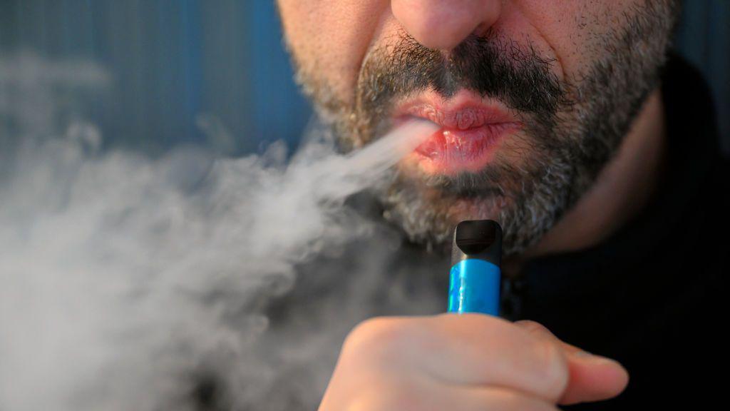 Man blowing out a puff of smoke from a vape. His lips are pursed and he has a dark black and grey speckled beard. He holds a blue vape clasped in his hand. 