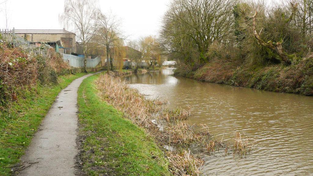 A grassy footpath runs beside a canal, which is brown in colour.