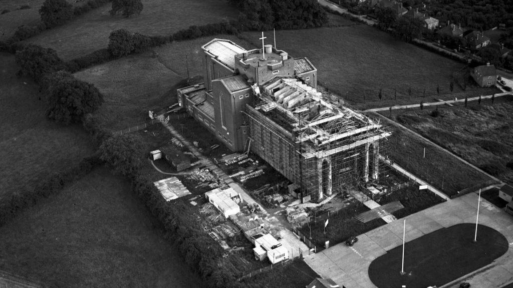 A brick cathedral under construction, mostly complete but with part of the roof missing and scaffolding on the exterior. The building is seen from above and in black and white.