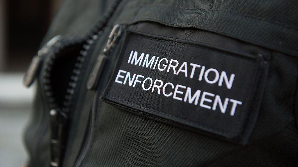 A close-up shot of a black immigration enforcement vest. The vest has 'IMMIGRATION ENFORCEMENT' written on it in white writing.