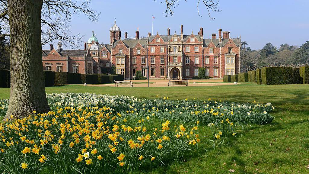 A view of The Church of St Mary Magdalene on Sandringham estate. Daffordils, a lush lawn and a tree are in the foregrounf of the large red-bricked building.