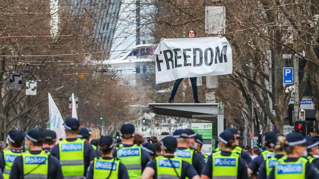 A man holds a banner reading "Freedom" atop a tram stop during an anti-lockdown protest in Victoria in 2021