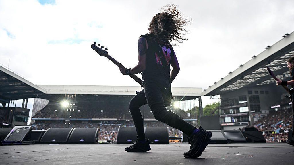 A man with a guitar and long hair plays on a stage, in front of a large crowd.
