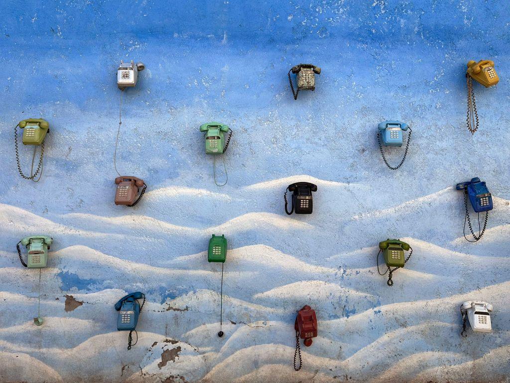 Dial-up landline telephones of various colours are mounted on a sky-blue wall.