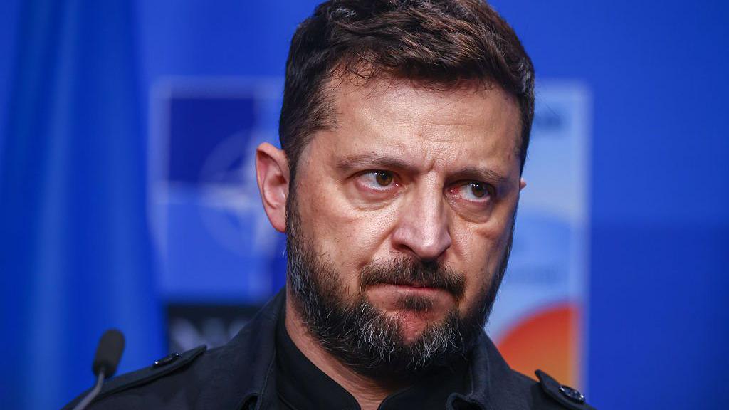 Ukrainian President Volodymr Zelensky sitting in front of a bright blue background. It is a close-up photo and his eyes are looking off to the right. He is sitting in front of a small microphone and the top of his black shirt is visible. 