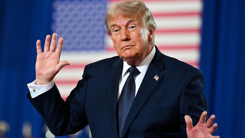 Trump holds up his hands while speaking at an event. He is wearing a dark suit and standing in front of an American flag